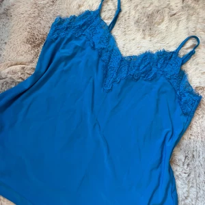 Blå linnetopp med spets från Amisu - Supersnygg blå linnetopp från Amisu med tunna axelband och v-ringning dekorerad med spets. Toppen är i ett mjukt, lätt material och har en enkel, stilren look som passar perfekt till jeans eller kjol. Perfekt för dig som gillar färg och detaljer!