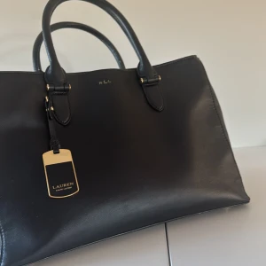 Svart handväska från Lauren Ralph Lauren - Stilren svart handväska i skinn från Lauren Ralph Lauren med guldfärgade detaljer och dubbla handtag. Väskan har en klassisk, fyrkantig form och dragkedja upptill. Insidan är fodrad i brunt tyg med logotyp och har flera fack för smart organisering.