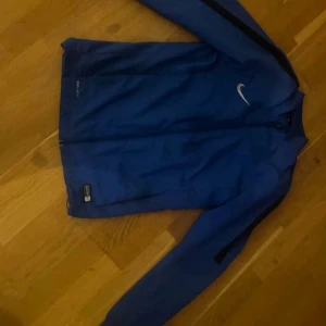 Blå Nike Dri-Fit vindjacka S - Blå vindjacka från Nike med Dri-Fit-teknologi. Jackan har hel dragkedja framtill, lång ärm och diskret Nike-logga på bröstet. Tillverkad i lätt polyester med meshfoder för extra ventilation. Perfekt för träning eller när du vill ha en sportig look.