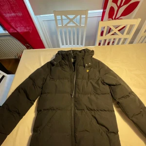 Svart pufferjacka från Lyle & Scott - Svart pufferjacka från Lyle & Scott med huva och dragkedja framtill. Jackan har en klassisk quiltad look och den ikoniska gula loggan på bröstet. Perfekt för kalla dagar och riktigt snygg till streetwear-stilen.