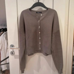 Brun kofta med knappar från Gina Tricot - Mysig brun kofta från Gina Tricot med rund halsringning och knäppning framtill. Modellen är lite croppad och har långa ärmar samt ribbade muddar. Perfekt att slänga över ett linne eller t-shirt för en chill look.