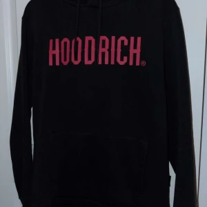 Svart HOODRICH hoodie med röd logga - Svart hoodie från HOODRICH med stor röd logga framtill. Klassisk känguruficka, huva med snörning och långa ärmar. Perfekt för dig som gillar streetwear och vill ha en stilren men ändå iögonfallande tröja.