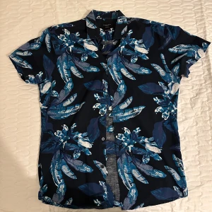 Blå blommig skjorta Selected Homme - Snygg kortärmad skjorta från Selected Homme med blått och vitt blommönster. Slim fit-modell i bomull och linne, perfekt för sommardagar. Skjortan har klassisk krage och knäppning framtill.