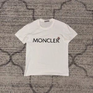 Vit Moncler t-shirt med logotyp - Vit t-shirt från Moncler i mjuk bomull med klassisk rund hals och korta ärmar. Stor Moncler-logga tryckt i svart på bröstet och en liten broderad Moncler-emblem bredvid. Perfekt för dig som gillar stilrena och exklusiva plagg.