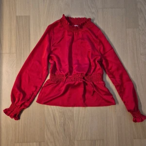 Röd blus med smock och volang - Snygg röd blus med smockdetaljer vid hals, midja och ärmslut. Blusen har långa ärmar och en liten volangkant vid halsen och ärmsluten. Perfekt för dig som vill ha en färgstark och trendig topp.