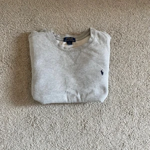 Grå sweatshirt från Polo Ralph Lauren - Klassisk grå sweatshirt från Polo Ralph Lauren med broderad logga på bröstet. Tröjan har rund halsringning, ribbade muddar och är tillverkad i mjuk bomullsmix. Perfekt för en chill och stilren look.