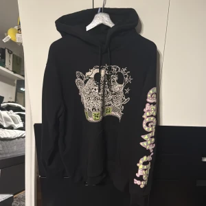 Svart hoodie MCQ - Svart hoodie från Alexander Mcqueen med coolt grafiskt tryck framtill och färgglad text längs ena ärmen. Tröjan har huva med snörning och är tillverkad i mjuk bomull. Perfekt för dig som gillar streetwear och unika detaljer.