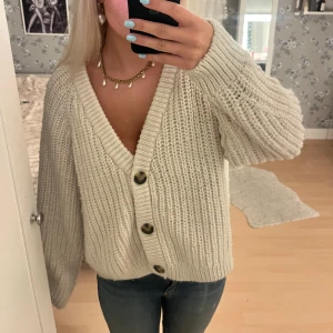 Vit stickad kofta med stora knappar - Supermysig vit stickad kofta med stora, snygga knappar framtill och djup v-ringning. Koftan har en loose passform och långa ärmar, perfekt att slänga på sig när det är kyligt. Passar grymt till jeans eller kjol för en chill look.