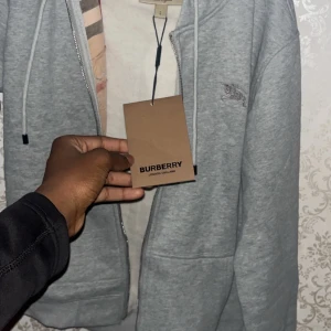 Grå hoodie från Burberry med dragkedja - Snygg grå hoodie från Burberry med dragkedja och klassiskt rutigt foder i huvan. Broderad logga på bröstet och snörning i huvan. Perfekt för dig som vill ha en stilren och exklusiv look.