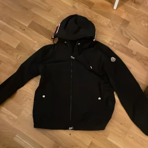 Monclerjacka - Svart vindjacka från Moncler med huva och dragkedja framtill. Jackan har två fickor med knappar och Moncler-logga på ärmen. Huvan har en snygg röd, vit och blå rand. Perfekt för dig som vill ha en stilren och sportig look.