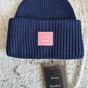 Mörkblå Acne Studios mössa i ull - Ribbstickad mörkblå mössa från Acne Studios i 100% ull. Klassisk modell med bred uppvikt kant och ikonisk rosa face-patch framtill. Perfekt accessoar för att hålla värmen och ge din outfit en cool touch.