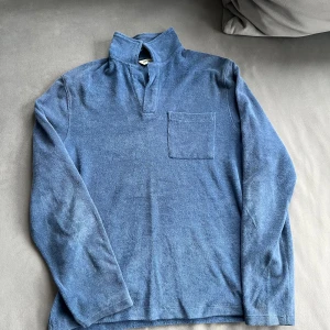 Blå långärmad tröja med krage - Säljer en blå långärmad tröja med hög krage och bröstficka. Tröjan är i mjukt frottématerial och har en avslappnad passform, perfekt för svala dagar. Enkel och stilren design som funkar till det mesta.