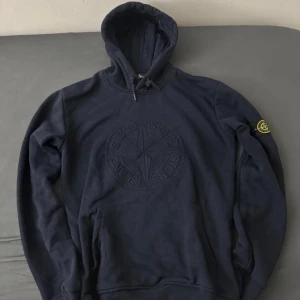 Mörkblå Stone Island hoodie M - Mörkblå hoodie från Stone Island i storlek M med broderad logga på bröstet och klassisk patch på ärmen. Tröjan har huva med snörning och känguruficka framtill. Perfekt för dig som gillar streetwear och vill ha en stilren look.