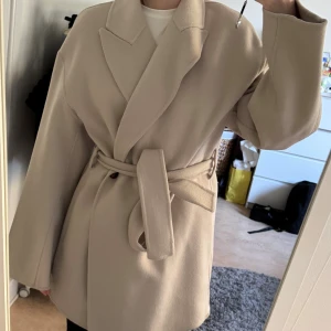 Beige ullmix kappa  - Stilren beige kappa med bred krage, knäppning framtill och matchande bälte i midjan. 50% ull. Använd fåtal gånger. Storlek xs passar även s. Nypris 1599:-