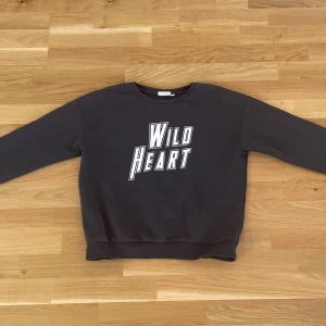 Svart sweatshirt Wild Heart KappAhl - Cool svart sweatshirt från KappAhl med trycket 'Wild Heart' i vitt framtill. Tröjan har rund hals, långa ärmar och ribbade muddar vid ärmslut och nederkant. Perfekt för dig som gillar en avslappnad och trendig stil. Jag använder XS/S och passar därför perfekt för de som använder det också 