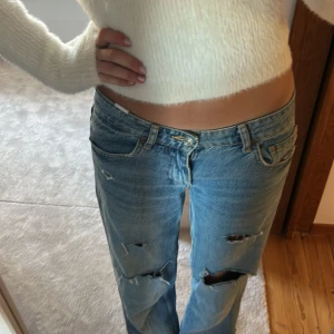 Blå slitna straight jeans - Säljer ett par blå jeans med raka ben och flera slitningar och hål på framsidan. Jeansen har klassisk femficksdesign, låg midja och är tillverkade i denim. Perfekta för en avslappnad och trendig look.