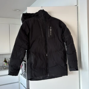 Svart pufferjacka från Sail Racing - Svart pufferjacka från Sail Racing med huva och flera dragkedjeförsedda fickor. Jackan har en stilren look med diskret logga på bröstet och bak på huvan. Perfekt för kalla dagar med sin vadderade design och långa ärmar.