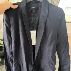 Svart kavaj med bälte från Lindex - Snygg svart kavaj från Lindex i storlek 36. Kavajen har klassisk krage, två fickor fram och ett matchande bälte i midjan för extra stilpoäng. Materialet är mjukt och har en lätt glansig finish, vilket ger en modern vibe. 