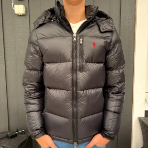 Svart Polo Ralph Lauren Dunjacka Glansig - Svart jacka från Polo Ralph Lauren med röd logga på bröstet. Jackan har huva, tvåvägsdragkedja och en bröstficka med dragkedja. Materialet är glansigt och väldigt varm. Den är endast använd fåtal gånger och är som ny. Inga defekter.