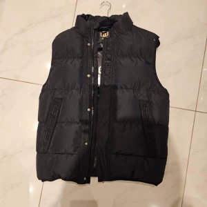 Svart dunväst från Glorious Gangsta - Svart dunväst med hög krage och dragkedja, dekorerad med broderad logga och mönsterdetaljer på både fram- och baksida. Insidan har ett grafiskt mönster i grått och svart. Västen har guldfärgade knappar och en cool streetstil.