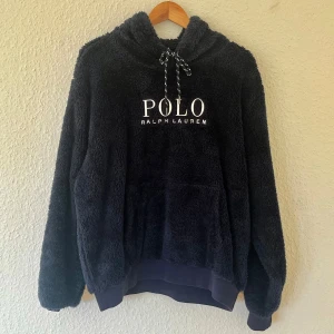 Marinblå teddyfleece - Polo Ralph Lauren - Mysig marinblå hoodie från Polo Ralph Lauren i fluffig fleece med broderad logga framtill. Tröjan har huva med snörning, känguruficka och långa ärmar. Perfekt för kalla dagar när du vill vara både bekväm och snygg. Passar även L.