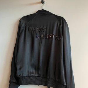 Svart bomberjacka från Saint Laurent - Svart bomberjacka från Saint Laurent med broderad logga på ryggen och detaljer vid fickorna. Jackan har ribbade muddar, dragkedja framtill och en clean, stilren look. Perfekt för dig som vill sticka ut med exklusiva detaljer.