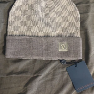 Louis Vuitton rutig grå mössa - Exklusiv mössa från Louis Vuitton i grått och beige med klassiskt rutigt mönster. Mössan har uppvikt kant med LV-logga framtill och är tillverkad i mjukt material som känns lyxigt. Perfekt accessoar för att lyfta din vinterstil.