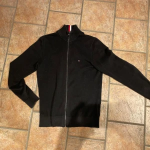 Zip tröja från Tommy Hilfiger  - Svart stickad kofta från Tommy Hilfiger med hel dragkedja och ståkrage med snygga rödvita detaljer. Liten logga på bröstet och ribbade muddar. Perfekt för dig som gillar klassisk stil med en modern touch.