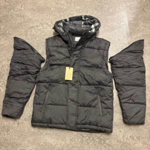 Svart pufferjacka från Burberry M - Svart pufferjacka från Burberry med avtagbara ärmar, huva fodrad med klassiskt rutigt tyg och dragkedja framtill. Jackan har två fickor med dragkedja och quiltad design. Perfekt för kalla dagar och riktigt snygg streetstil.