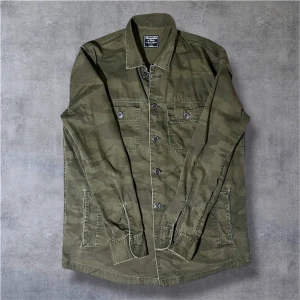 Camouflage overshirt från Abercrombie - Snygg overshirt från Abercrombie & Fitch i grönt camouflage-mönster. Skjortan har klassisk krage, två bröstfickor med knappar och långa ärmar. Tillverkad i mjuk bomull med detaljerade sömmar och knappstängning framtill. Perfekt för en cool streetstil. Kan tänka mig gå ner i pris vid snabb affär! Ha de bra! // Gidlöfs