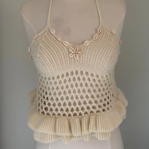Stickad beige linnetopp med snäckor - Unik stickad linnetopp i beige med virkade detaljer och volangkant. Toppen har knyt i nacken och är dekorerad med små snäckor längs urringningen. Perfekt för sommarens alla äventyr och ger en bohemisk vibe.