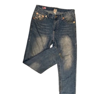 True Religion  - Säljer ett par klassiska blå jeans från True Religion med tydliga vita kontrastsömmar och ikoniska ficklock bak. Modellen har normal passform och raka ben, med snygga detaljer på fickorna. Perfekt för dig som gillar street och premium denim.