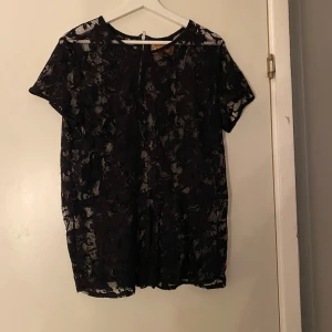 Svart spetsblus med dragkedja - Svart blus i genombruten spets med blommigt mönster och korta ärmar. Toppen har rund halsringning och en synlig dragkedja bak. Perfekt för dig som gillar detaljer och vill ha en snygg och stilren look.