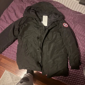 Ganada goose chateau parka - Säljer en svart jacka från Canada Goose i storlek L. Jackan har huva, dragkedja och knappar framtill samt den klassiska loggan på ärmen. Fodrad och riktigt varm, perfekt för kalla dagar. Snygg och stilren design som funkar till allt.