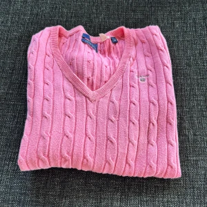 Rosa kabelstickad tröja från GANT - Supersnygg rosa kabelstickad tröja från GANT med v-ringning och broderad logga på bröstet. Tröjan har lång ärm och klassisk passform. Tillverkad i mjuk bomull som känns skön mot huden. Använd men i bra skick. Storlek M