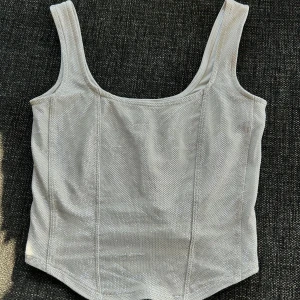 Glittrig silver topp från H&M Divided - Säljer en snygg glittrig silverfärgad korsett från H&M Divided. Toppen har breda axelband, croppad passform och markerade sömmar framtill. Materialet är en mix av polyester, metallfiber och elastan som ger en stretchig känsla och coolt skimmer. Använd endast en gång!! 