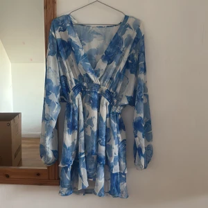 Blåvit blommig kortklänning med v-ringning - Superfin kortklänning med blått och vitt blommönster, v-ringad och med långa, luftiga ärmar. Klänningen har markerad midja med smock och volangig kjol. Tunt, flowy material som känns perfekt för varmare dagar.