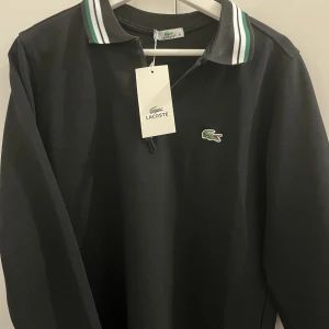 Svart pikétröja från Lacoste - Svart långärmad pikétröja från Lacoste med klassisk krage och grönt-vitt randigt mönster på kragen. Tröjan har dragkedja vid halsen och den ikoniska krokodilloggan broderad på bröstet. Perfekt för en clean och sportig look.