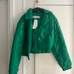 Grön quiltad pufferjacka från Bershka - Säljer en grön, croppad pufferjacka från Bershka i storlek XS. Jackan är quiltad med stora rutor, har en bred krage, tryckknappar och resår i midjan. Materialet är syntet och jackan har en riktigt trendig vibe.