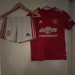 Manchester United fotbollströja Adidas - Röd Manchester United fotbollströja från Adidas med vita och röda detaljer, klubbmärke på bröstet och Chevrolet-logga framtill. Kortärmad modell i lätt funktionsmaterial, perfekt för match eller träning.