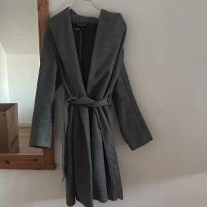 Grå kappa med huva från Zara - Snygg grå kappa från Zara med stor huva och knytskärp i midjan. Kappan har lång ärm och en rak, lite loose passform. Perfekt för dig som vill ha en stilren och enkel look med extra edge tack vare huvan.
