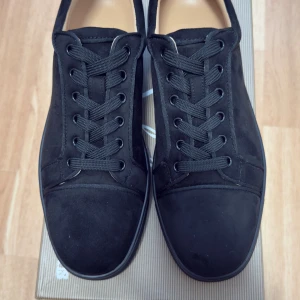 Svarta sneakers från Christian Louboutin - Snygga svarta sneakers från Christian Louboutin i mocka med klassisk röd sula och broderad logga på hälen. Skorna har rund tå, platt sula och snörning framtill. Perfekt för dig som gillar exklusiv streetstyle och vill sticka ut med detaljer.