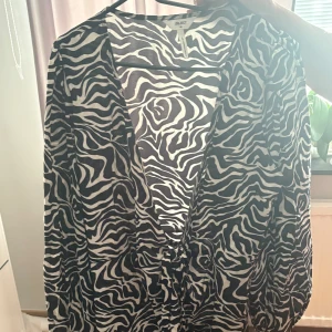 Zebramönstrad klänning från Object - Snygg zebramönstrad klänning från Object med svartvita vågiga ränder. Klänningen har v-ringad hals, lång ärm och knyt i midjan för en cool siluett. Perfekt för dig som vill sticka ut med ett djurinspirerat mönster.