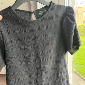 Svart blus med puffärm från SHEIN - Svart blus från SHEIN med puffiga korta ärmar och rund hals. Blusen har ett strukturerat, ribbat tyg med subtilt mönster och knapp i nacken. Perfekt för dig som gillar stilrena och trendiga detaljer.