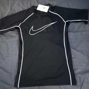 Nike Pro svart träningströja - Nike Pro svart träningströja med vit logga och kontrastsömmar. Tight passform och korta ärmar, perfekt för intensiva träningspass. Tillverkad i stretchigt syntetmaterial som andas och ger bra rörelsefrihet. Passar S bättre 