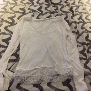 Vit långärmad blus med spetsdetaljer - Säljer en vit långärmad blus med v-ringning och fina spetsdetaljer både vid halsen och längs nederkanten. Blusen har en mjuk och lätt ribbad struktur som gör den extra skön att bära. Perfekt för dig som gillar romantiska och feminina detaljer.