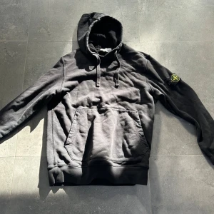 Svart hoodie från Stone Island - Svart hoodie från Stone Island med klassisk patch på ärmen. Tröjan har huva med snörning och en stor magficka framtill. Mjuk bomullskvalitet och långärmad modell, perfekt för en avslappnad streetwear-look.