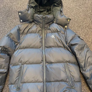 Blå pufferjacka från Calvin Klein - Blå pufferjacka från Calvin Klein med avtagbar huva och dragkedja framtill. Jackan har två snedställda fickor med dragkedja och tydliga CK-loggor på bröstet och ärmen. Perfekt för kalla dagar med sin vadderade design. Storlek XS men passar även perfekt för dem som har storlek S i vanliga fall.