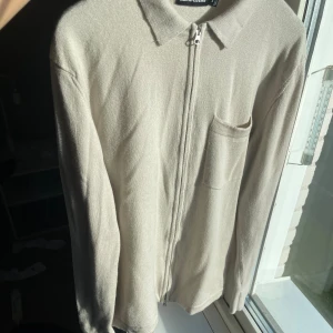 J.Lindeberg tröja  - VIKTIGT! Tröjan är storlek M men passar storlek S. Snygg beige kofta från J.Lindeberg med dragkedja framtill, klassisk krage och en bröstficka. Stickad i mjukt material och har lång ärm. Perfekt för lager-på-lager och stilren look.