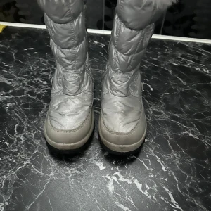 Silvriga moon boots med snöflingor - Säljer ett par silvriga moon boots med quiltad design och broderade snöflingor på sidorna. Skorna har en platt sula med svart och beige mönster undertill, samt rund tå. Perfekta för vintern och håller fötterna varma.